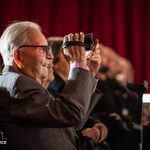 Starszy mężczyzna w garniturze robi zdjęcie aparatem na sali z czerwoną kurtyną w tle.