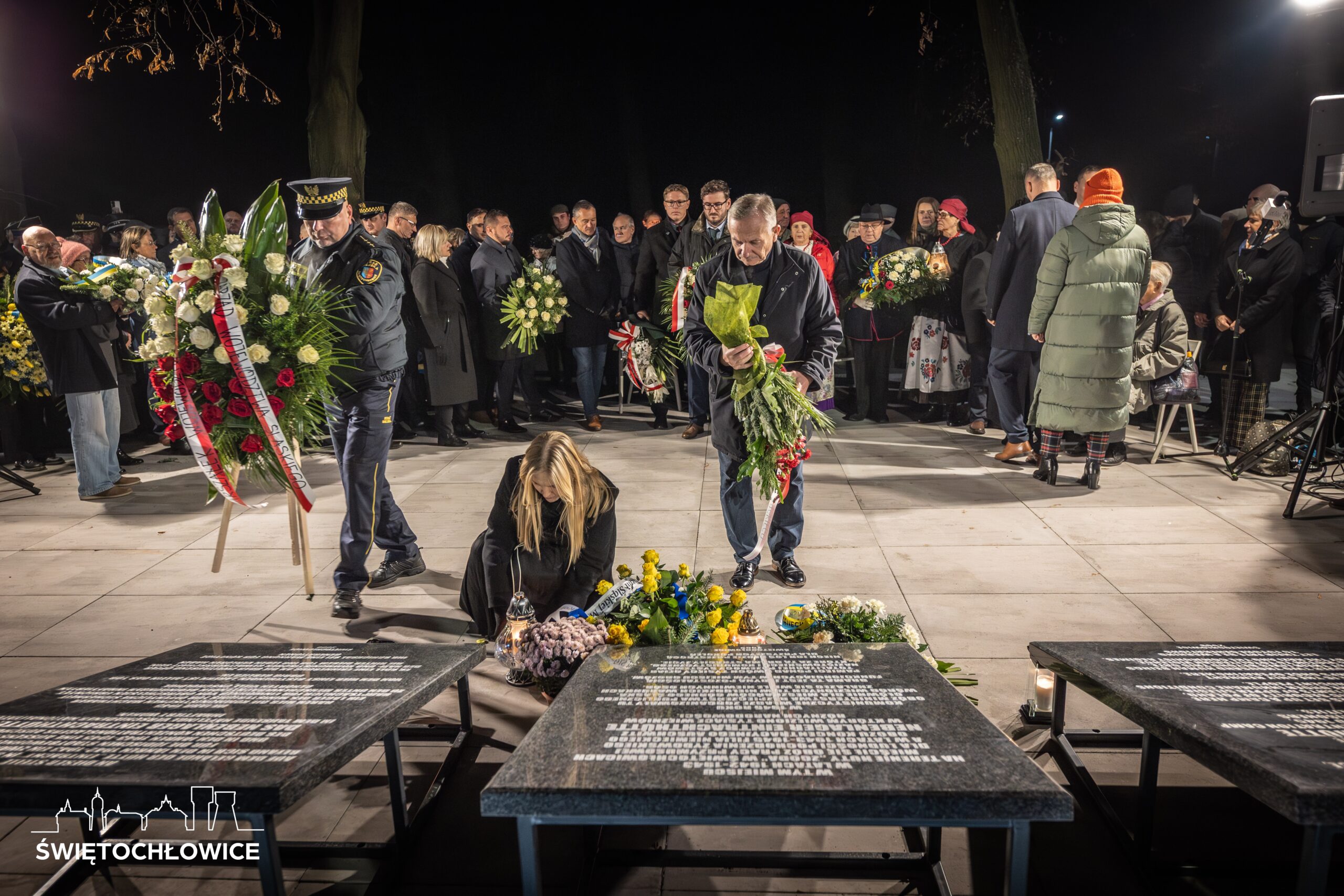 Uczestnicy uroczystości składają kwiaty pod tablicami pamiątkowymi przy tłumie zebranych.