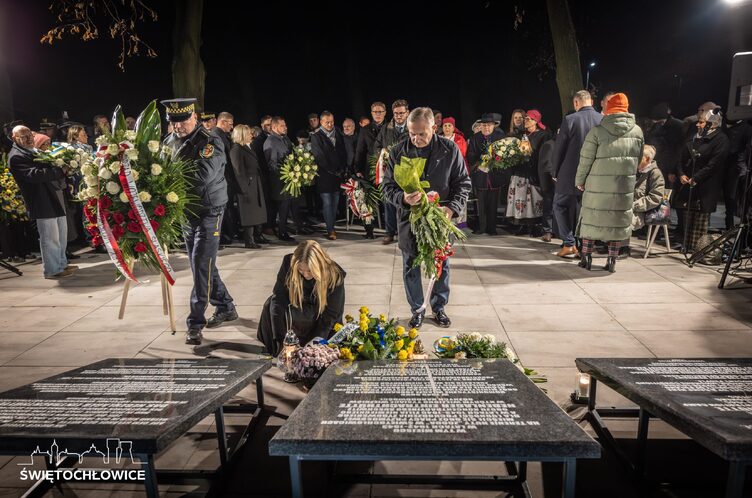 Uczestnicy uroczystości składają kwiaty pod tablicami pamiątkowymi przy tłumie zebranych.