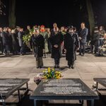 Grupa osób w garniturach składa hołd pod tablicami pamiątkowymi nocą, w tle zgromadzenie.