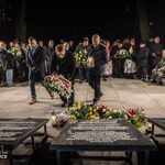 Dwóch mężczyzn z wieńcem i lampionem składa hołd przy tablicy pamiątkowej, w tle uczestnicy
