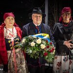 Trzy osoby w strojach ludowych, kobiety z chustami i mężczyzna z wieńcem, podczas uroczystości.