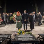 Trzy osoby stojące przy tablicy pamiątkowej, wokół zgromadzeni, składanie hołdu.