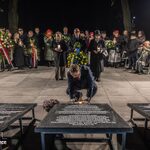 Mężczyzna klęka przy tablicy i zniczu podczas uroczystości wieczornej, wokół tłum.